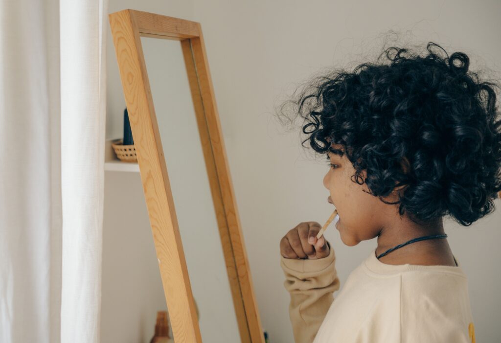 Side view of focused African American child with curly hair standing opposite mirror and brushing teeth while looking at mirror in bathroom